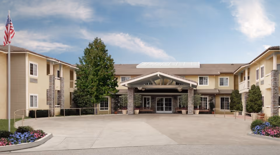 Front exterior view of a two-story senior living facility building with a covered entrance supported by stone pillars, surrounded by landscaping including trees and flower beds, and an American flag on a flagpole to the left.