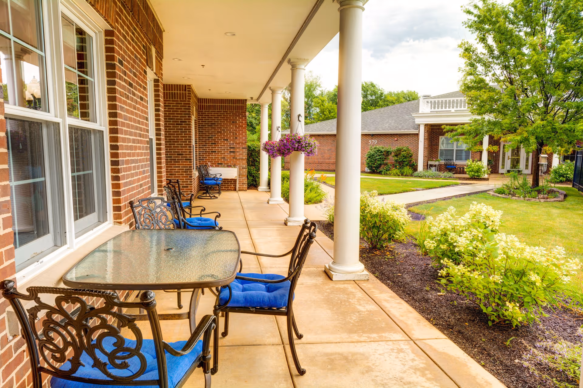Covered outdoor patio area with a glass-top table and ornate metal chairs with blue cushions. The patio is supported by white columns and overlooks a well-maintained garden with green shrubs and a tree. In the background, there is a brick building with windows and a walkway leading through the garden.