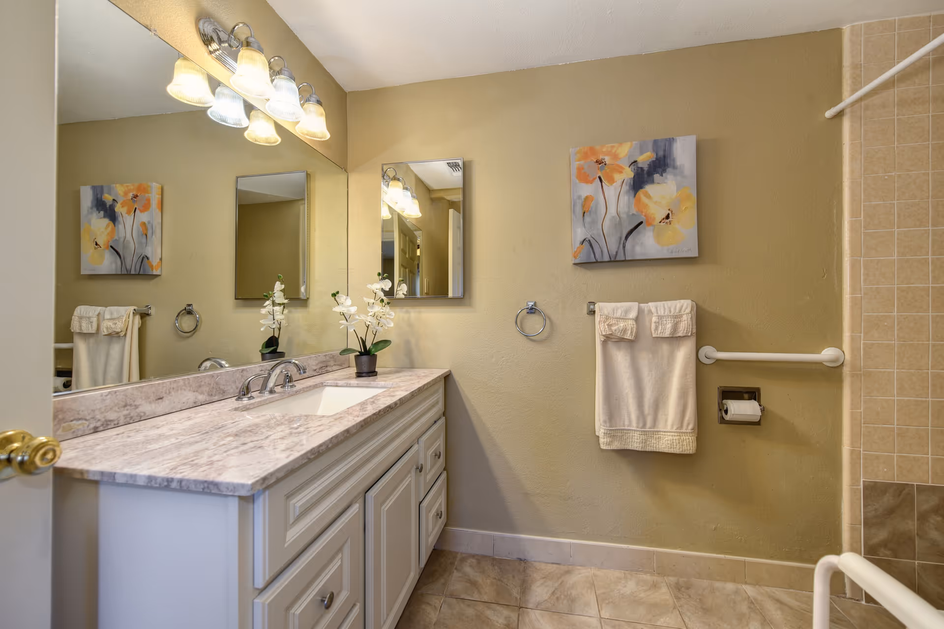 A bathroom with a beige wall and tiled floor. It features a white vanity with a marble countertop and an under-mount sink. Above the sink is a large mirror with a light fixture containing four lamps. There is a small potted white orchid on the countertop. On the wall, there is a towel rack with a beige towel and a floral painting with yellow and orange flowers. A grab bar and a toilet paper holder are visible next to a tiled shower area.