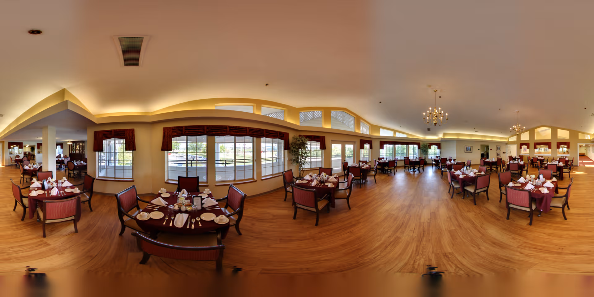 A spacious dining room with multiple round tables set with white napkins, plates, and silverware. The room has large windows with red valances allowing natural light to fill the space. The floor is wooden, and there are chandeliers hanging from the ceiling. The overall atmosphere is warm and inviting.