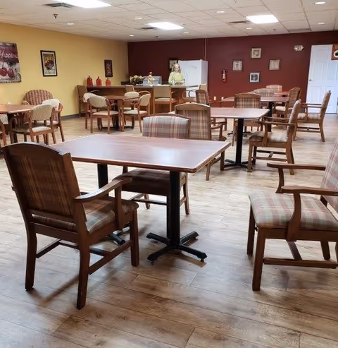 A spacious dining area with multiple wooden tables and cushioned chairs arranged neatly on a wooden floor. The walls are painted in warm tones, with framed pictures and decorative items. In the background, a person stands near a counter with a refrigerator and other kitchen items.