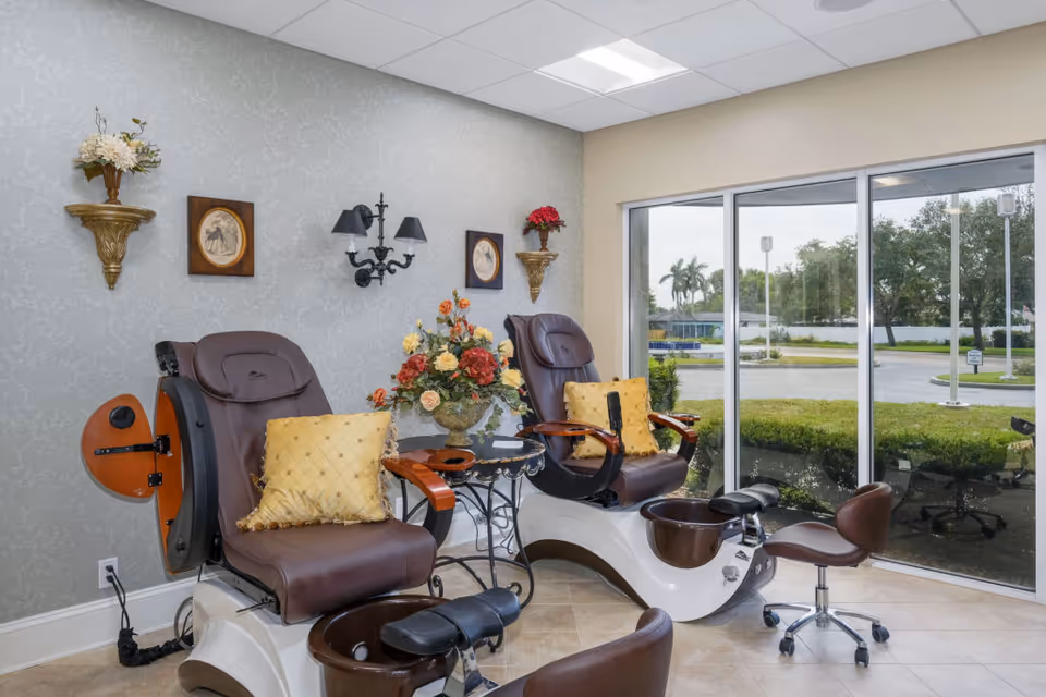 Two brown pedicure chairs with foot baths, decorative wall shelves and a floral centerpiece beside large windows overlooking the facility grounds.