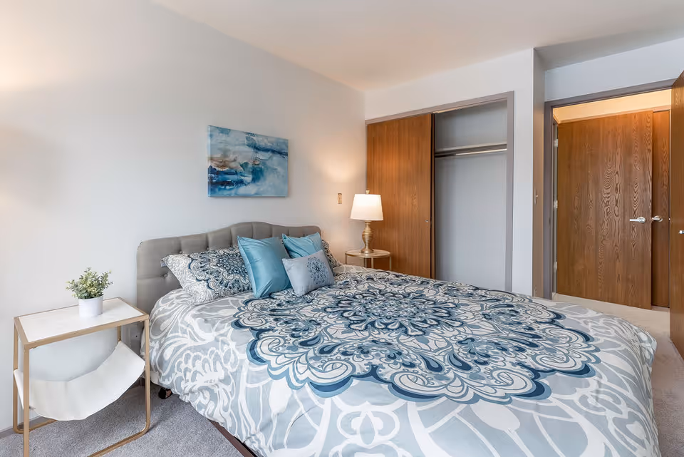 A bedroom with a bed featuring a blue and white patterned comforter and multiple pillows. There is a small side table with a potted plant on the left and a round table with a lamp on the right. A wooden closet with sliding doors is open, and a wooden door to the room is visible in the background. A blue abstract painting hangs on the wall above the bed.