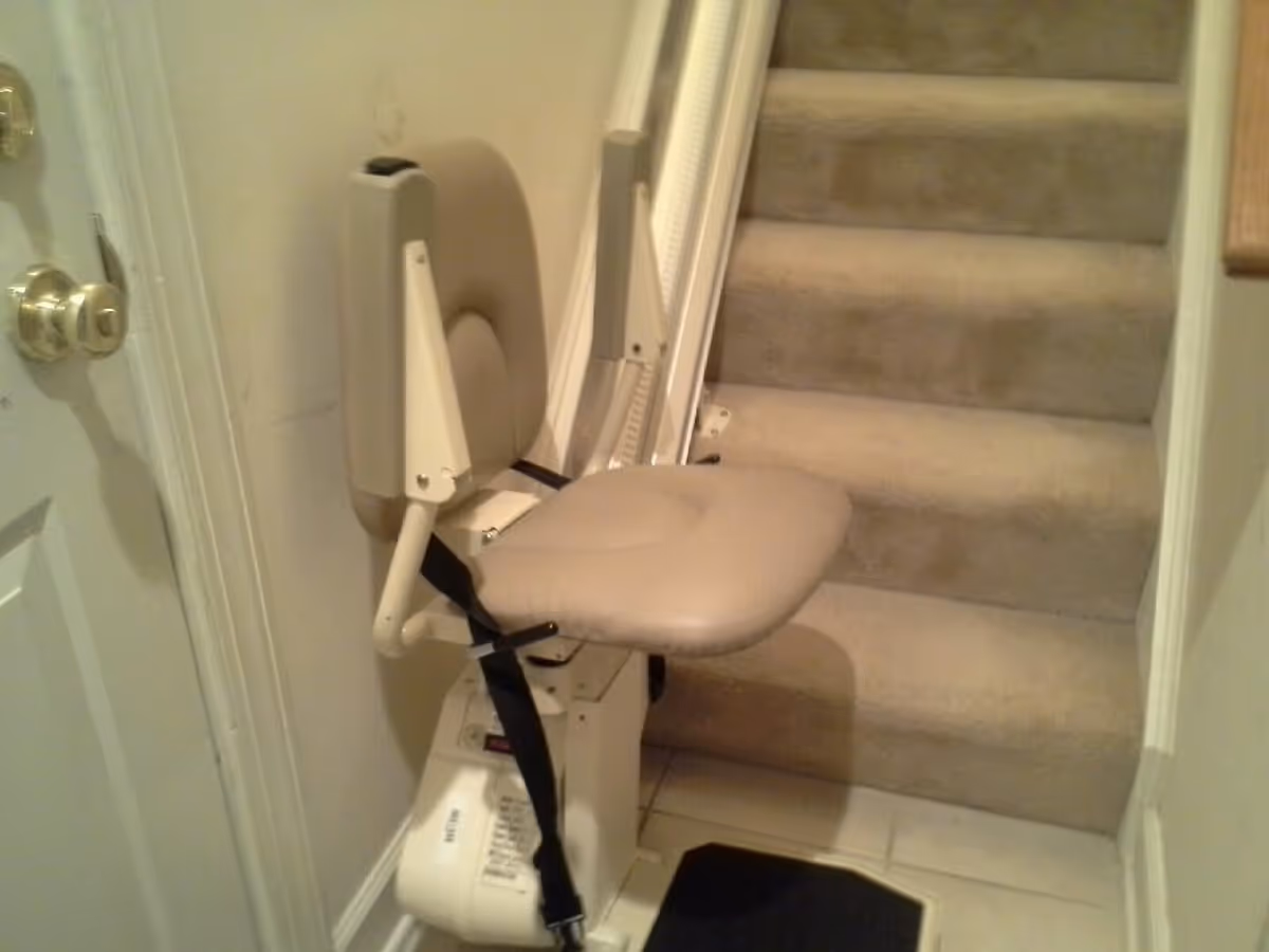 A beige stairlift installed on a carpeted staircase next to a white door with brass doorknobs. The stairlift has a padded seat and backrest with a safety belt.