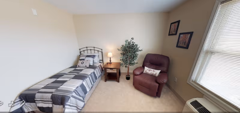 A small bedroom with a single bed covered in a gray and white checkered blanket, a wooden nightstand with a lamp, a potted plant, a maroon recliner chair with a decorative pillow, two framed pictures on the wall, and a window with blinds letting in natural light.