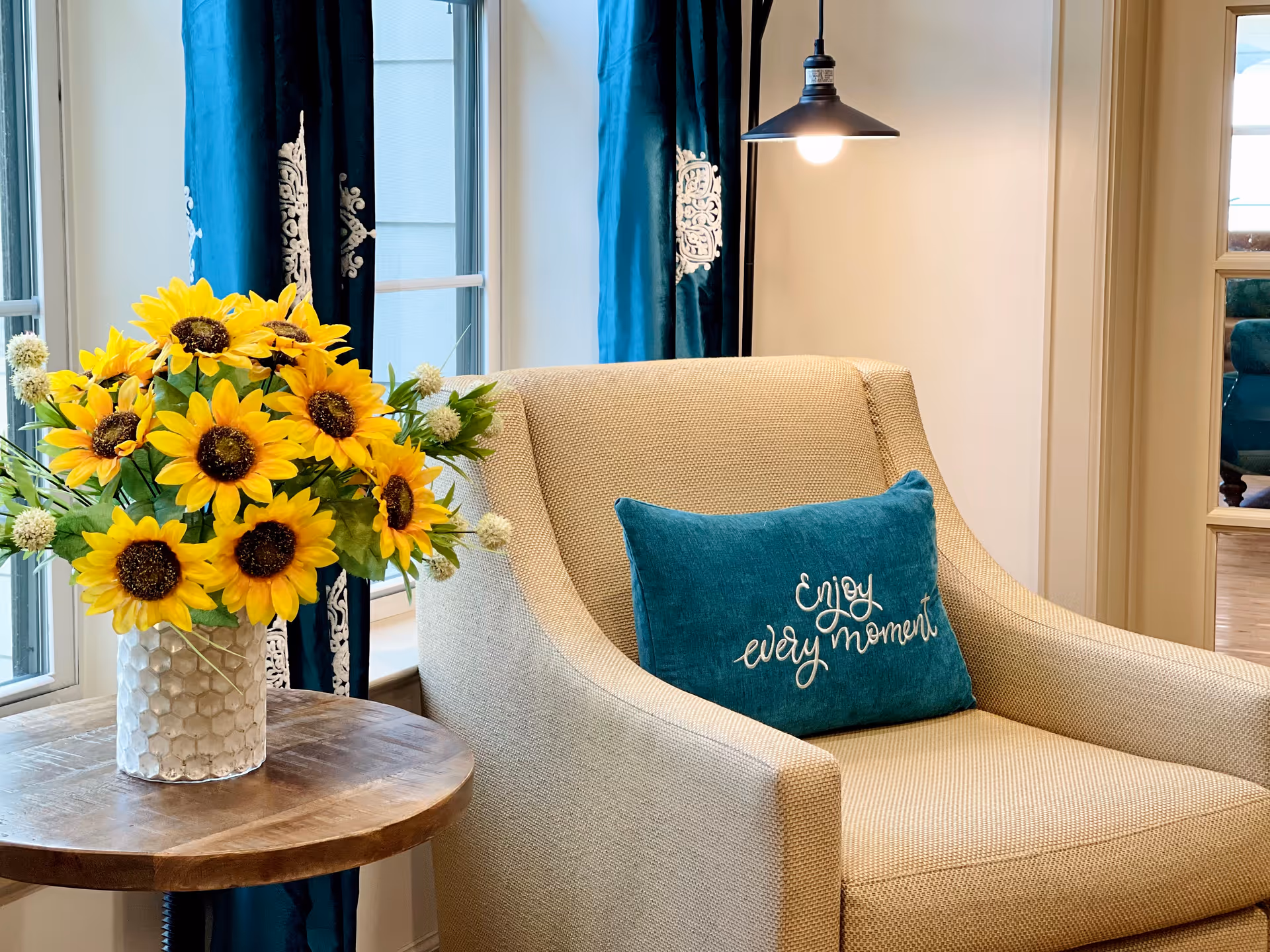A cozy beige armchair with a teal pillow that says 'Enjoy every moment' next to a wooden side table holding a vase of bright yellow sunflowers. Behind the chair are windows with blue curtains featuring white decorative patterns, and a hanging black pendant light above the chair.