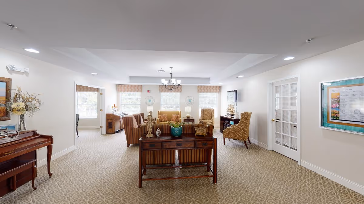 A bright and spacious senior living common area with beige patterned carpet, multiple armchairs and sofas arranged around a central wooden table, a piano on the left, and a TV mounted on the right wall. The room has large windows with valances, white walls, and a chandelier hanging from the ceiling.