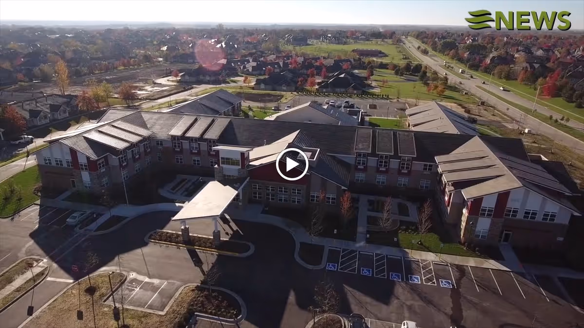 Aerial view of a large nursing home facility with multiple connected buildings, surrounded by parking lots and landscaped areas. The facility is situated in a suburban neighborhood with roads and houses visible in the background under a clear sky.