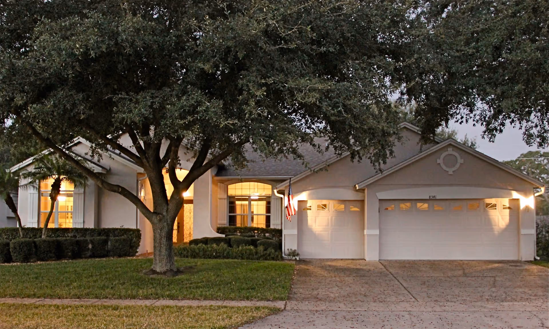 Exterior view of a single-story residential building with a large tree in the front yard, a well-maintained lawn, and a driveway leading to a double garage. The house has light-colored walls, multiple windows with shutters, and an American flag mounted near the garage. Warm interior lights are visible through the windows.