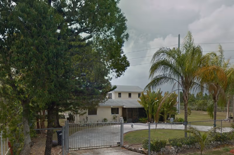 Front exterior view of a two-story residential building with a gated driveway, surrounded by trees and palm plants under a cloudy sky.