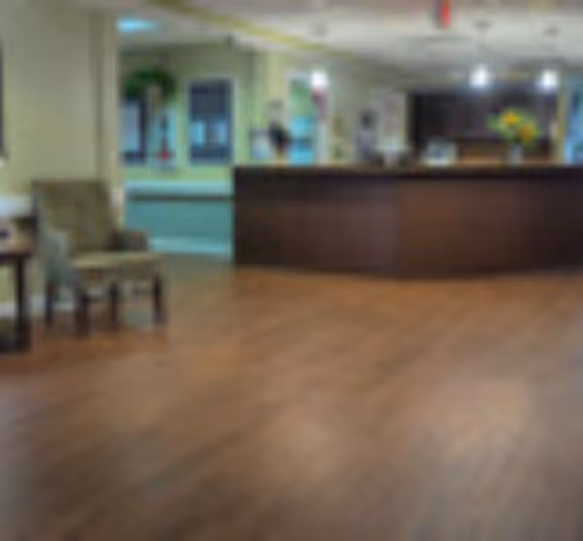 Interior view of a senior living facility reception area with a wooden floor, a dark wooden reception desk, chairs along the left wall, and a bright, well-lit background.