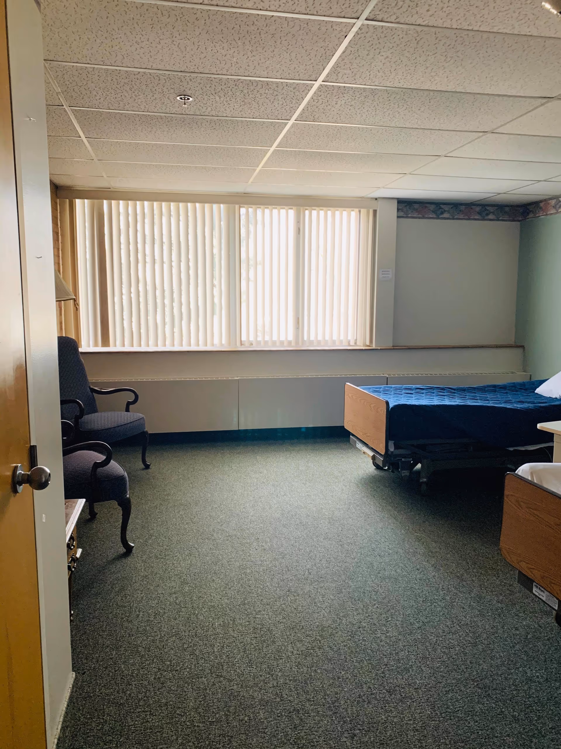 A simple bedroom with a hospital-style bed covered with a blue blanket, a pillow, two upholstered chairs, a small wooden table, and a large window with vertical blinds letting in natural light.