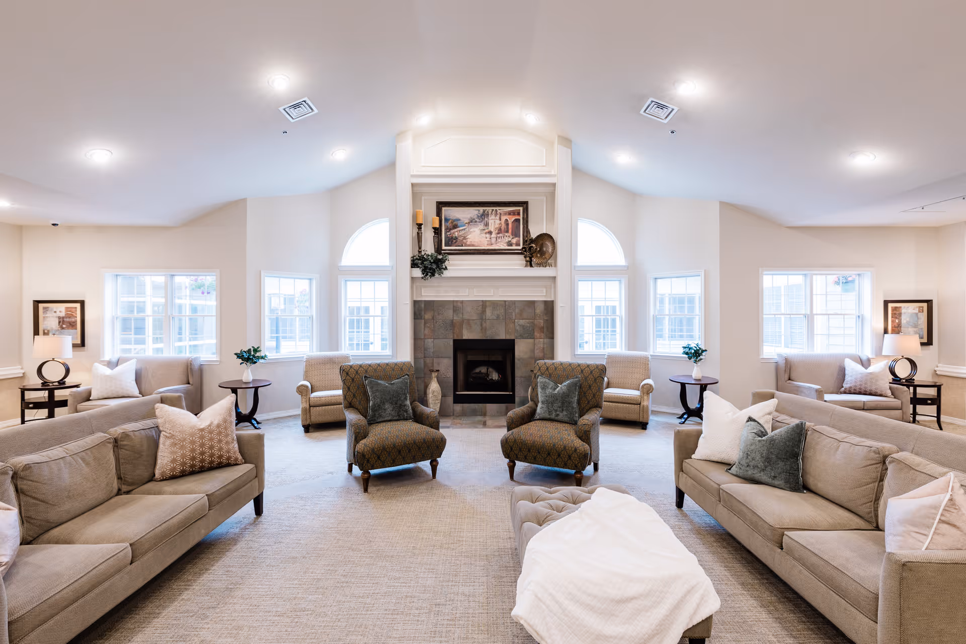 A spacious and well-lit living room in a senior living facility with two beige sofas, two patterned armchairs, and two beige armchairs arranged around a central fireplace. The room features multiple windows allowing natural light, side tables with lamps and plants, and a framed painting above the fireplace.
