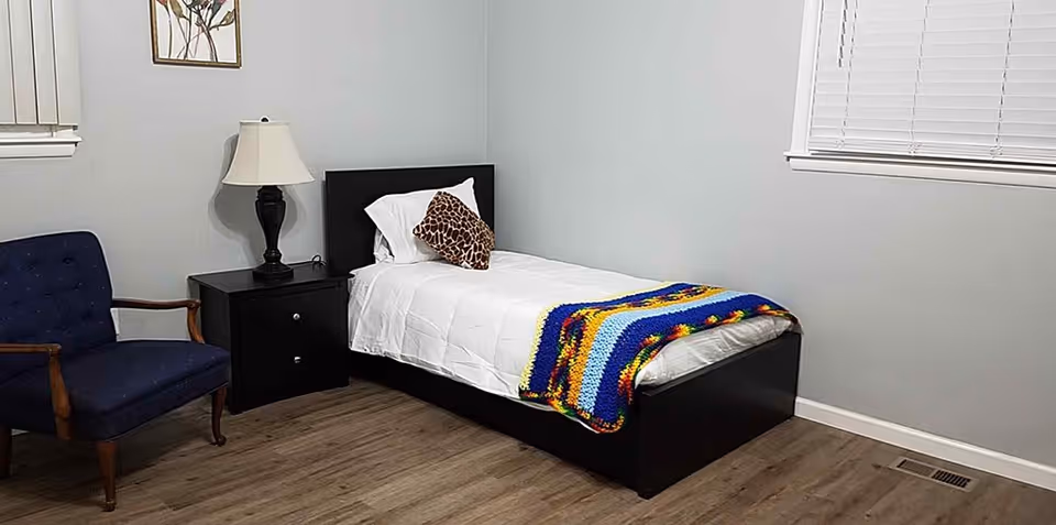 A simple bedroom with a single bed featuring white bedding, a colorful knitted blanket, and a giraffe-patterned pillow. Next to the bed is a black nightstand with a lamp. There is a blue upholstered chair with wooden armrests and legs. The room has light gray walls, two windows with white blinds, and wood flooring.