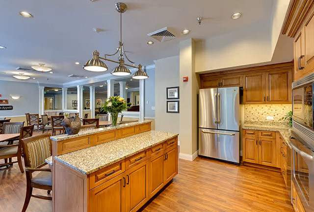 A spacious kitchen area with wooden cabinets, granite countertops, and stainless steel appliances including a refrigerator and built-in oven. The kitchen island has a vase with flowers and a decorative rooster figurine. In the background, there is a dining area with tables and chairs, and soft lighting from ceiling fixtures.