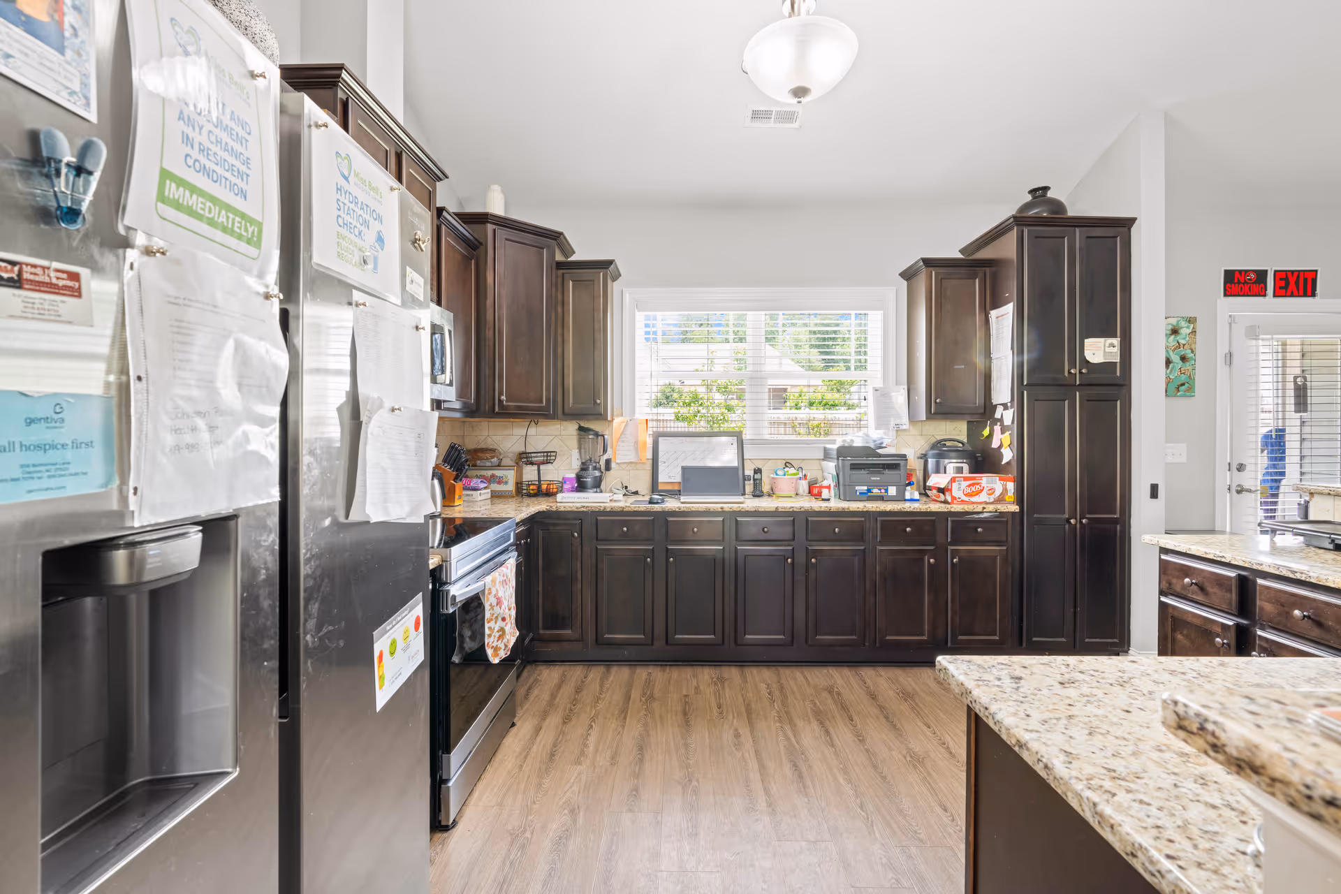 Bright, spacious kitchen with dark wood cabinets, stainless steel refrigerator and stove, granite countertops, and a window above the sink.