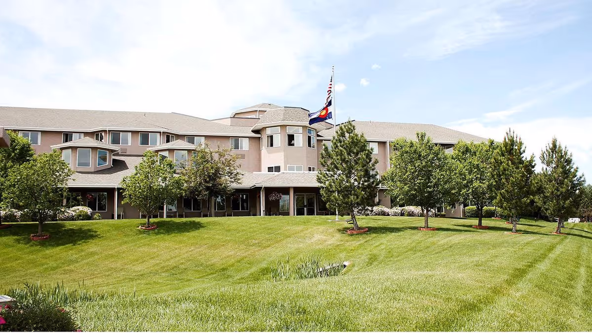 Front exterior of a multi-story senior living building with a manicured lawn, trees, and flagpoles.