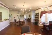 A spacious dining room with wooden floors and multiple round tables set with red napkins and white plates. The room features chandeliers hanging from the ceiling, large windows with curtains allowing natural light, and a serving counter on the left side. There are chairs around the tables and decorative shelving along the walls.