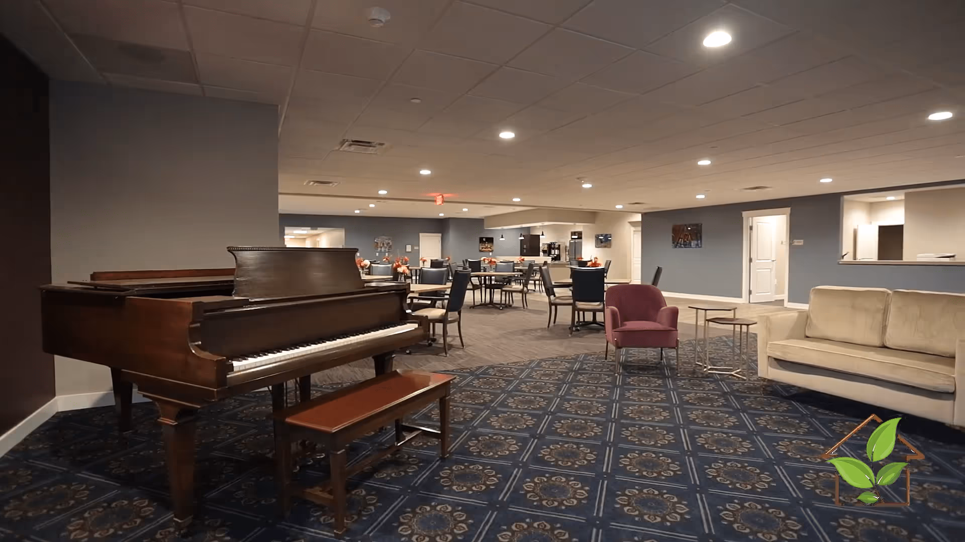 Interior of a senior living facility common area featuring a grand piano with a bench, several round tables with chairs, a purple armchair, and a beige sofa. The room has patterned blue carpet, blue and beige walls, and recessed ceiling lights. There is a serving window to a kitchen area in the background.