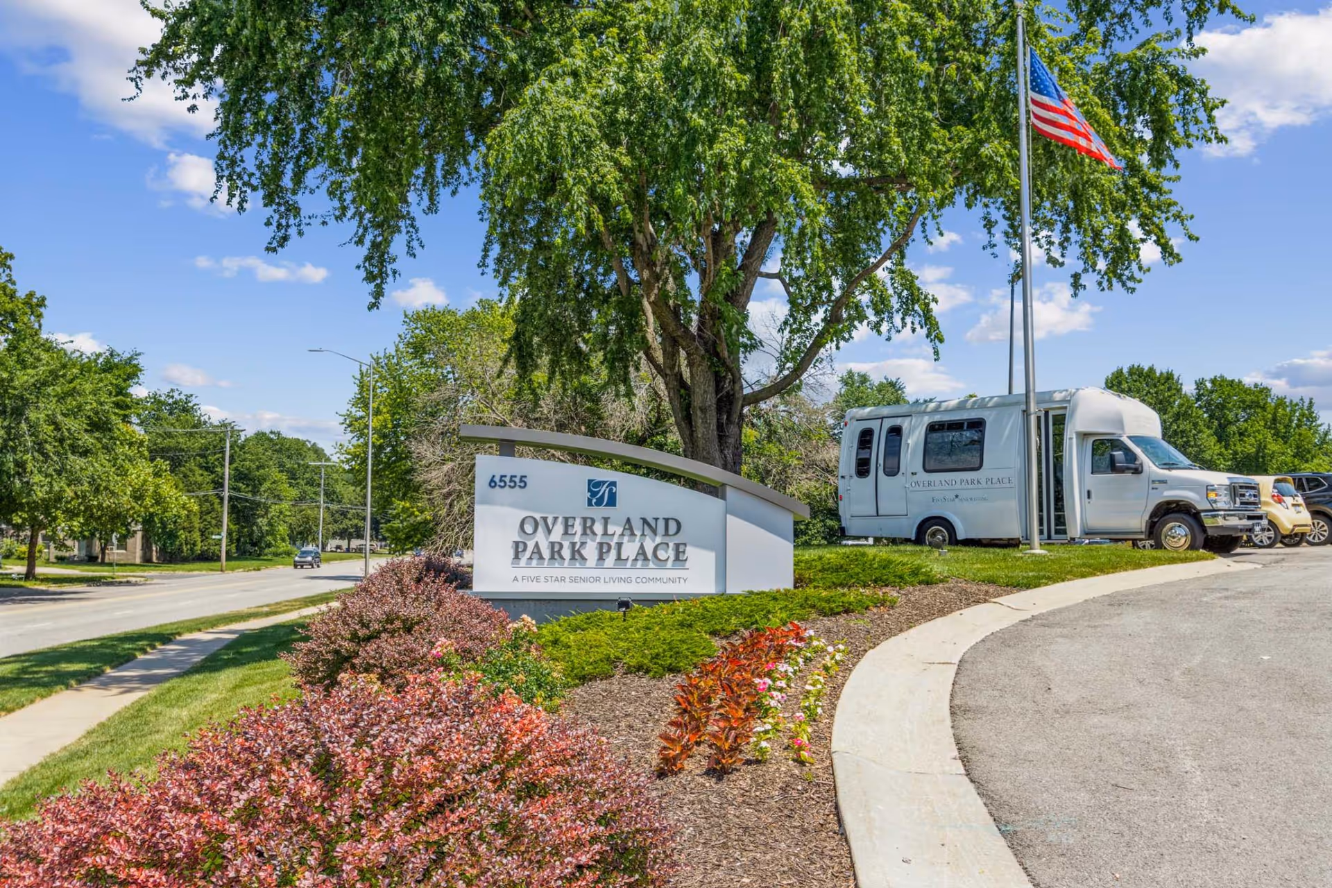 Outdoor view of the entrance to Overland Park Place senior living community, featuring a landscaped area with colorful bushes and flowers, a large tree, a sign displaying the facility name and address, an American flag on a pole, and a white shuttle van parked nearby.