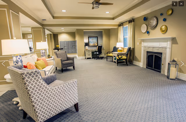 A spacious and well-lit living room area in an assisted living facility featuring patterned armchairs, a sofa with colorful cushions, a fireplace with decorative plates above it, a desk with a chair and computer, and large windows with curtains.