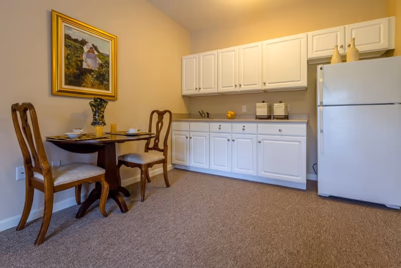 A small kitchen area with white cabinets, a white refrigerator, and a countertop with a sink. To the left, there is a small round wooden dining table set with two chairs, a vase with flowers, and tableware. A framed painting of a person in a field hangs on the wall above the table. The floor is carpeted.