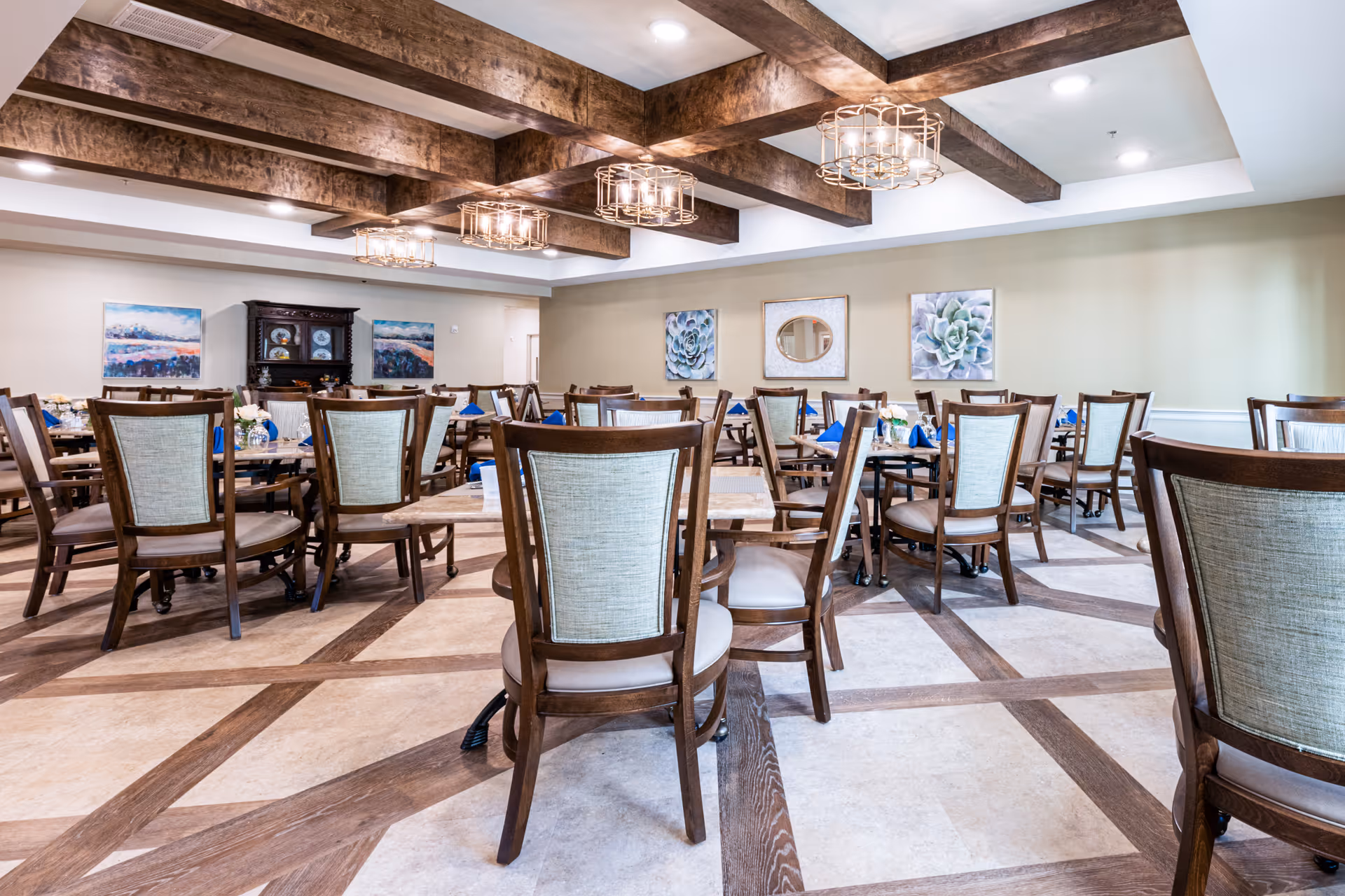 A spacious dining room with multiple wooden tables and chairs arranged neatly. The ceiling features exposed wooden beams with modern light fixtures. The walls are decorated with framed artwork and a round mirror. The floor has a patterned design with wood and tile elements.
