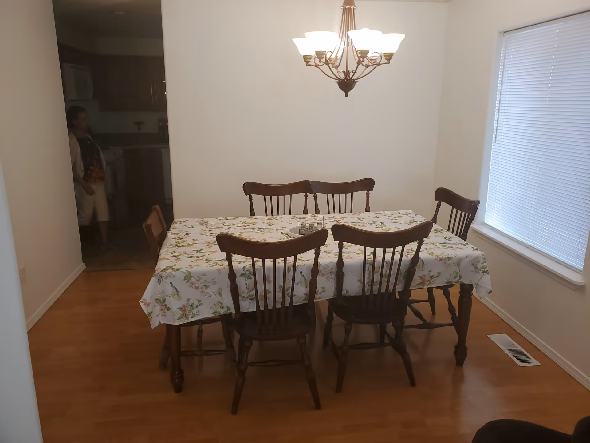 A dining room with a wooden table covered by a floral tablecloth and six wooden chairs around it. A chandelier with multiple lights hangs above the table. There is a window with closed blinds on the right wall, and a kitchen area is visible in the background with a person standing near the entrance.
