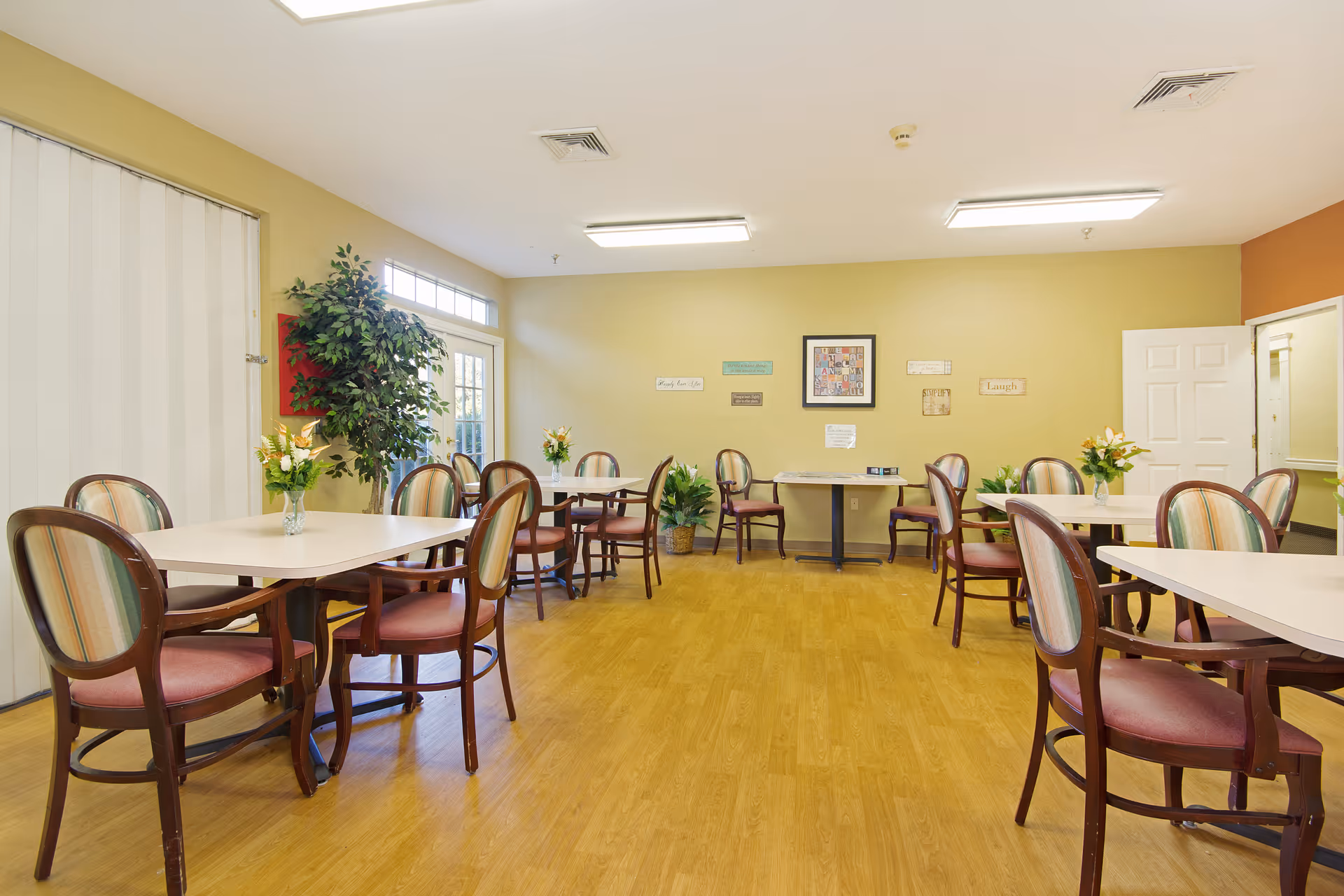 A bright dining room with several tables and chairs arranged neatly. Each table has a small vase with flowers. The room has light yellow walls, wooden flooring, and fluorescent ceiling lights. There are decorative plants and framed artwork on the walls.