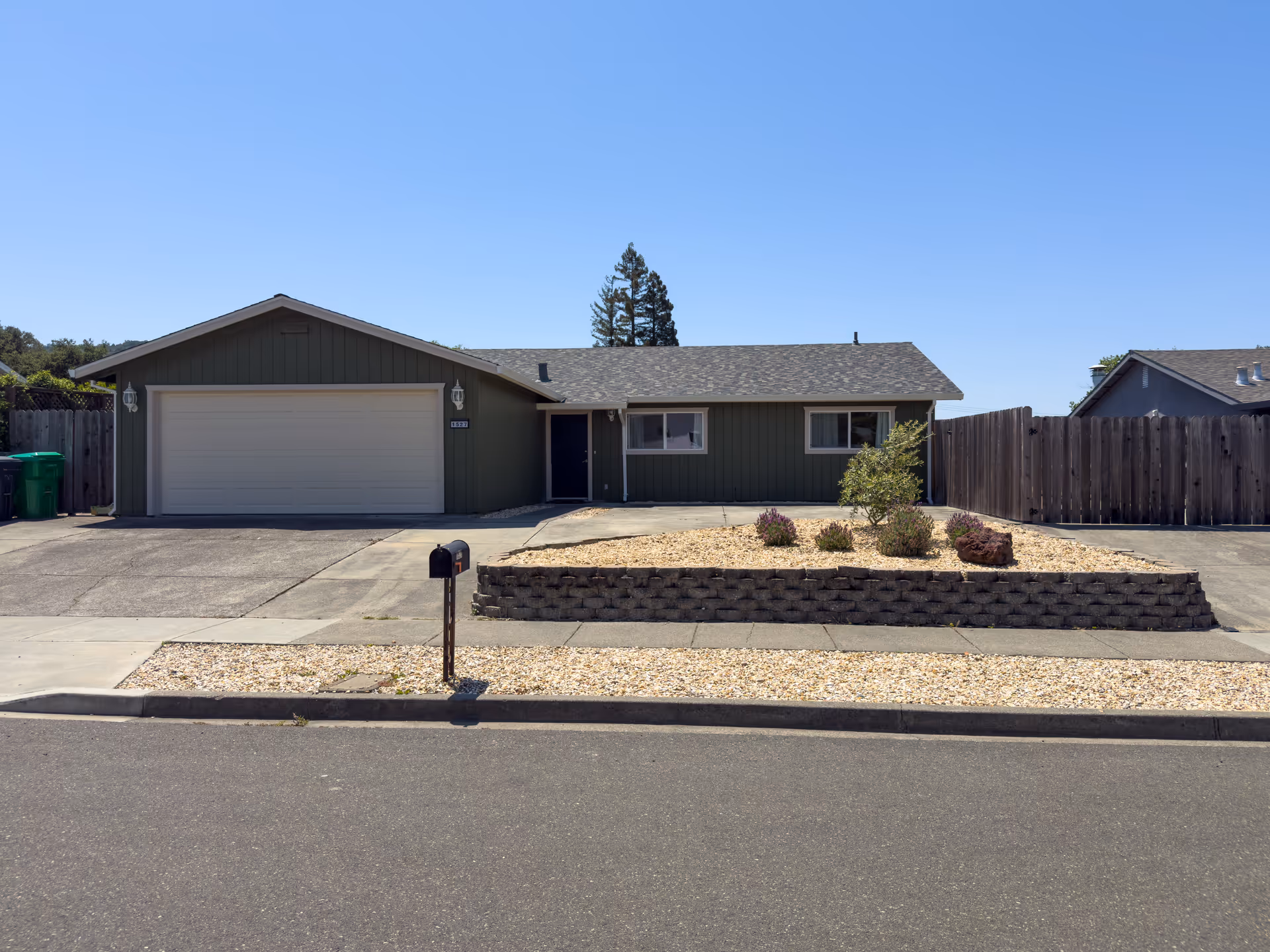 Single-story green house with an attached two-car garage, low rock landscaping, a mailbox and wooden fence under a clear blue sky.