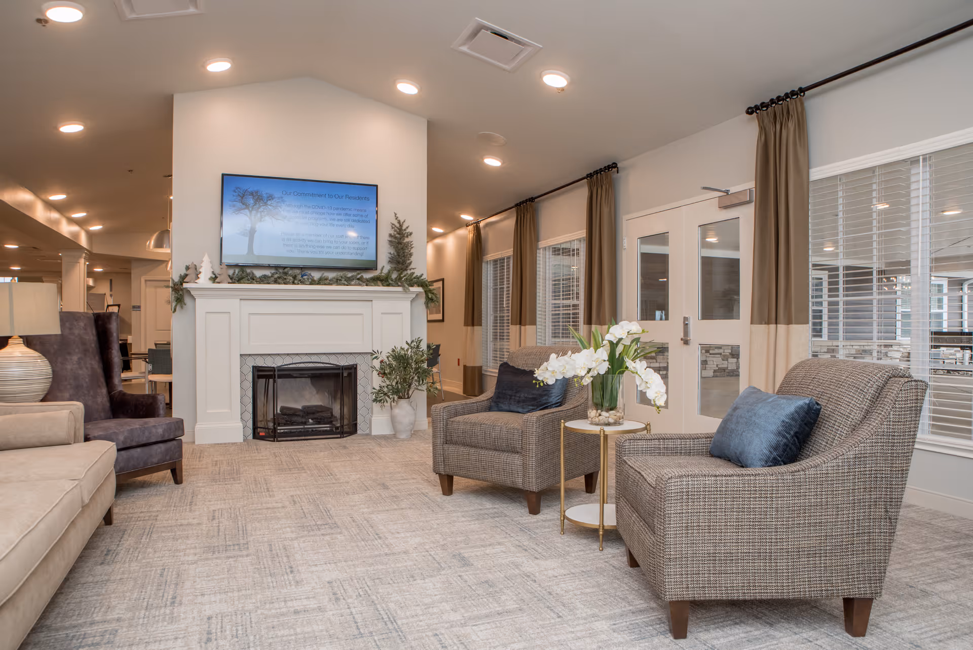 A cozy senior living facility common area with two cushioned armchairs with blue pillows, a small round table with a vase of white flowers, a fireplace with decorative greenery on the mantel, and a flat-screen TV mounted above the fireplace. The room has large windows with beige and brown curtains and light-colored carpeted flooring.