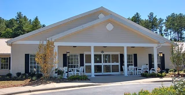 Exterior front view of a single-story building with a covered entrance supported by white columns. There are white chairs and small tables on the porch area, surrounded by small trees and shrubs, with a backdrop of tall pine trees.
