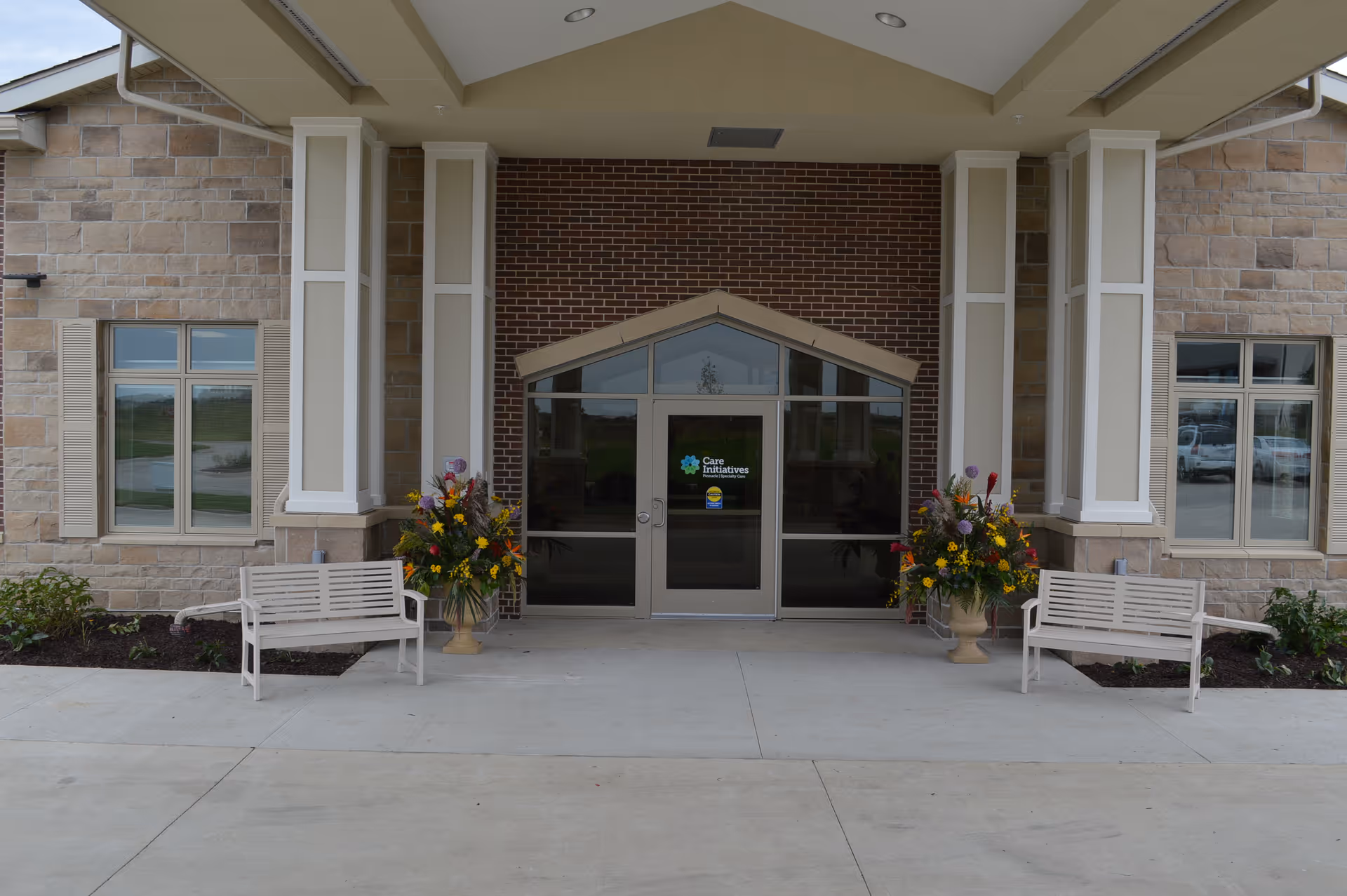 Entrance of a building with a covered porch supported by columns, two white benches on either side, large flower arrangements in urns, and glass double doors with a sign that reads 'Care Initiatives'. The building exterior features stone and brick walls with windows on both sides.