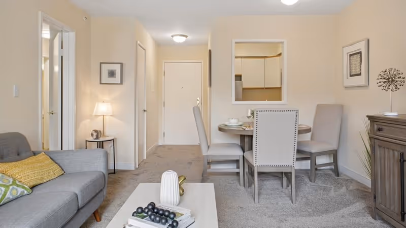 Open-plan living and dining area with a sofa, coffee table, round dining table with chairs, and the entry door visible.
