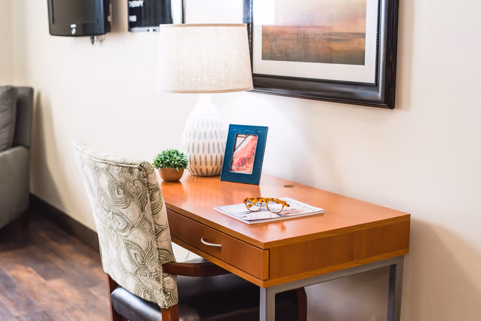 Wood desk with a table lamp, framed photo, small potted plant, glasses and a patterned chair in a cozy interior.