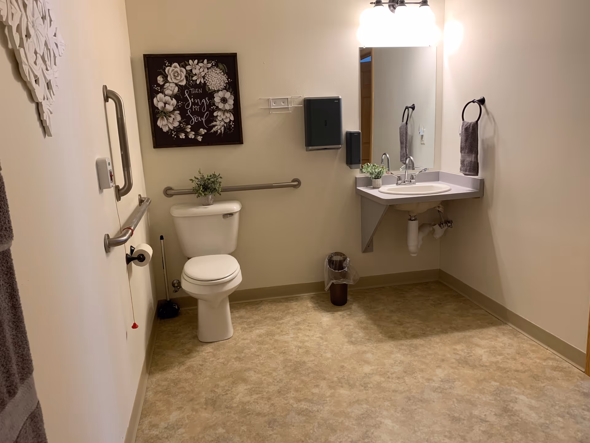 A clean and accessible bathroom with beige walls and flooring. The room features a white toilet with a small potted plant on top, grab bars on the walls for support, a wall-mounted sink with a mirror above it, a towel ring holding a gray towel, a small trash can, and a decorative framed floral artwork on the wall.