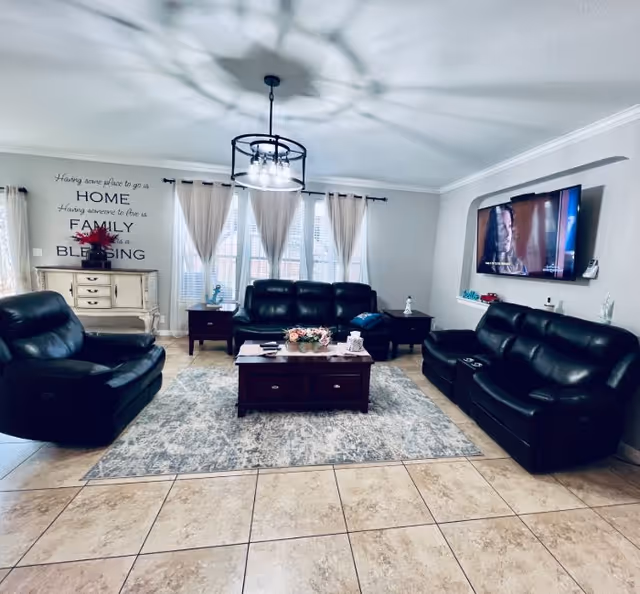 A cozy living room with three black leather sofas arranged around a wooden coffee table on a patterned rug. The room has tiled floors, large windows with sheer curtains, and a wall-mounted flat-screen TV. A decorative light fixture hangs from the ceiling, and a small cabinet with a red flower arrangement is against the wall with a quote about home, family, and blessing.