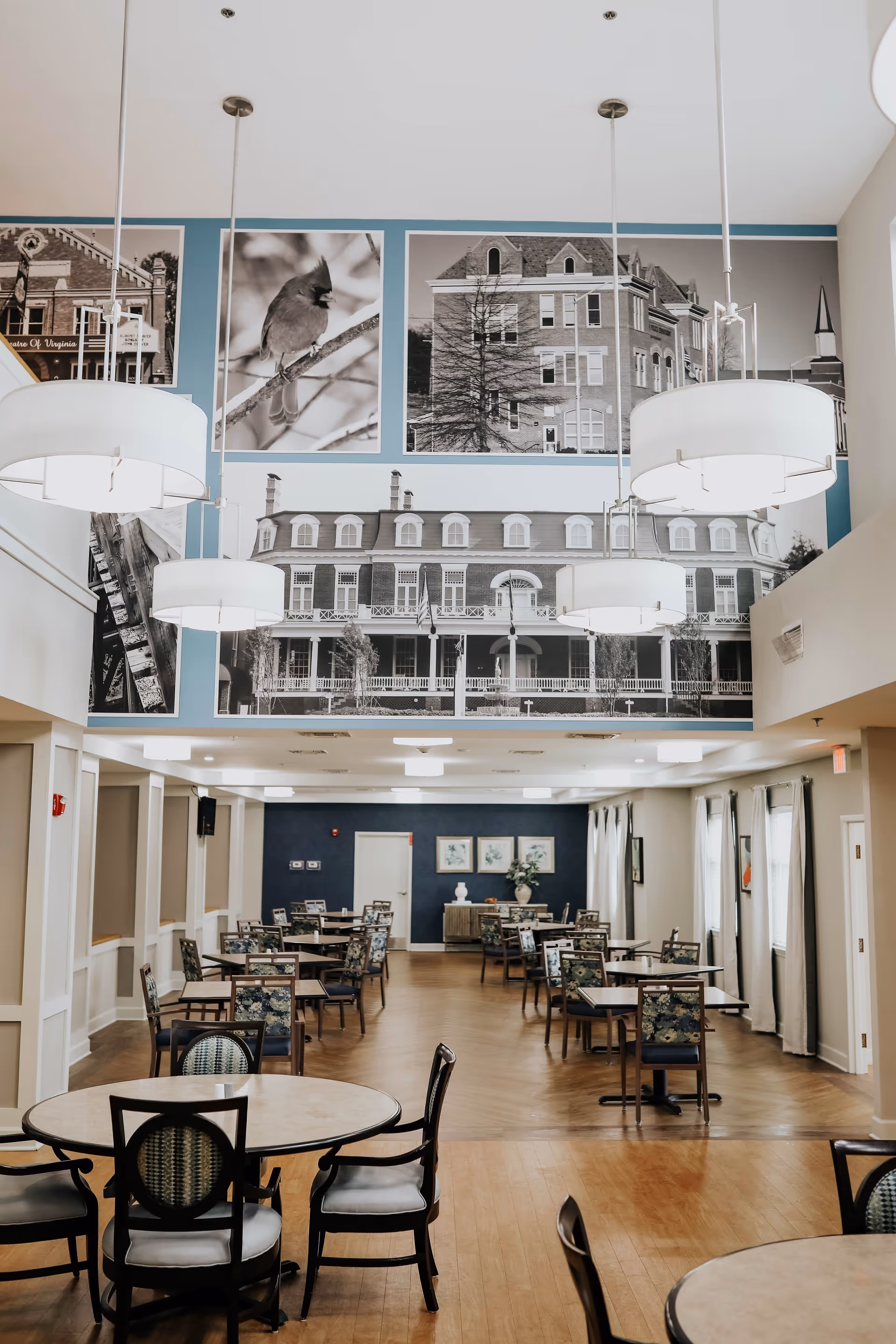 A spacious dining room with multiple round and square tables surrounded by chairs. The room features large hanging pendant lights and a wall decorated with large black and white photographs of buildings and a bird. The floor is wooden, and there are windows with curtains on the right side.
