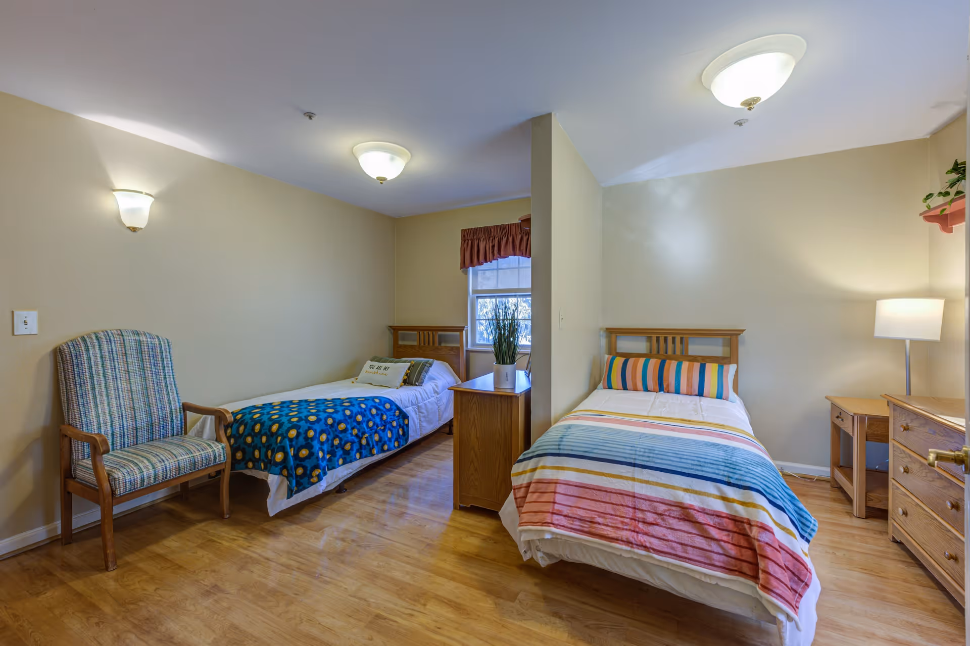 A shared bedroom in a senior living facility with two single beds separated by a partial wall. Each bed has colorful bedding, one with a blue blanket with yellow patterns and the other with a striped blanket in shades of blue, pink, and orange. There is a wooden chair with striped upholstery on the left side, a wooden dresser and desk with a lamp on the right side, and a window with a valance in the back. The room has wooden flooring and beige walls with ceiling and wall-mounted light fixtures.
