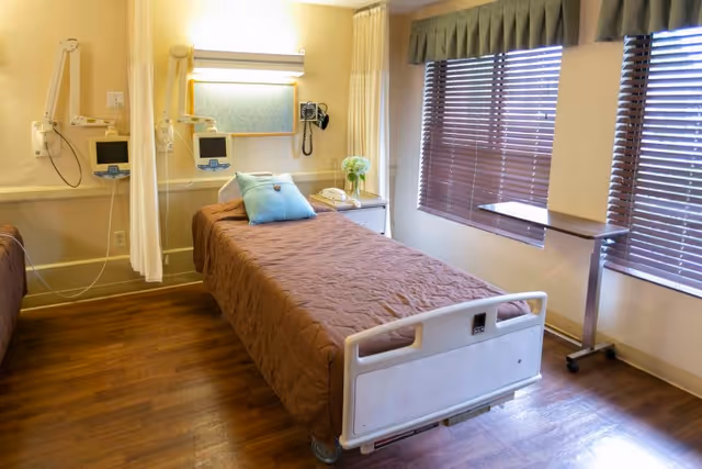 A single hospital-style bed with a brown coverlet in a bright patient room with medical monitors, a bedside table, and large windows with blinds.