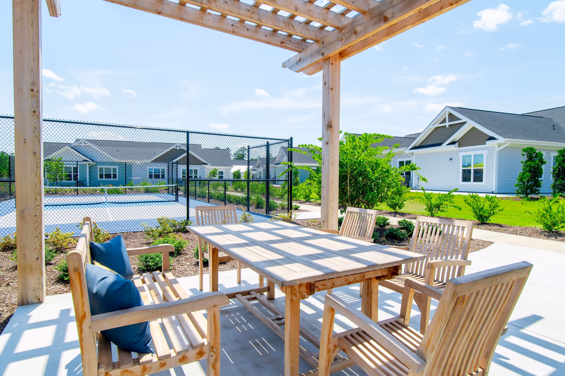 Outdoor seating area with a wooden table and chairs under a pergola, overlooking a fenced tennis court and landscaped greenery with residential buildings in the background under a clear blue sky.