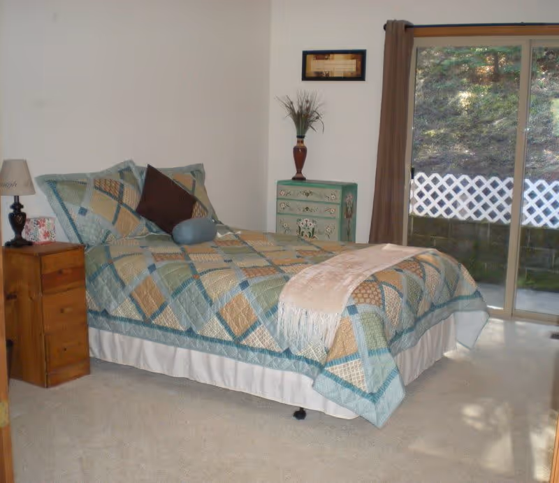 A neatly made bed with a patterned quilt in a bedroom with wooden nightstands, a small painted dresser, and a sliding glass door to the outside.