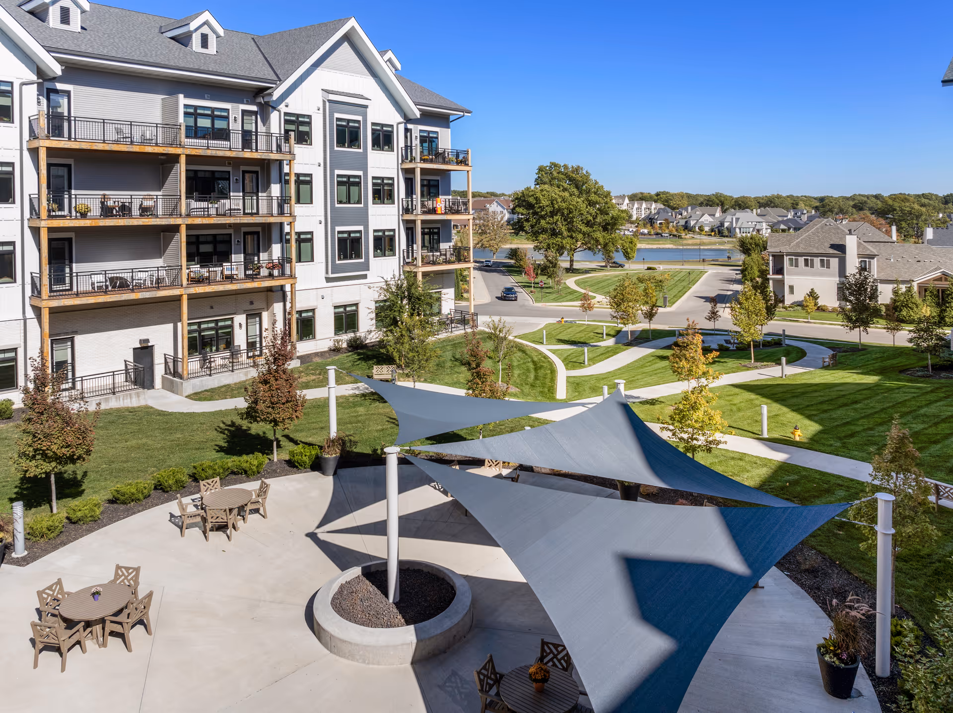Outdoor view of a senior living facility showing a multi-story building with balconies, a landscaped courtyard with green lawns, trees, walking paths, and shaded seating areas with tables and chairs under large fabric canopies. In the background, there are additional residential buildings and a body of water under a clear blue sky.