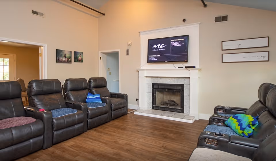 Communal living room with rows of leather recliners facing a fireplace and a wall-mounted TV.
