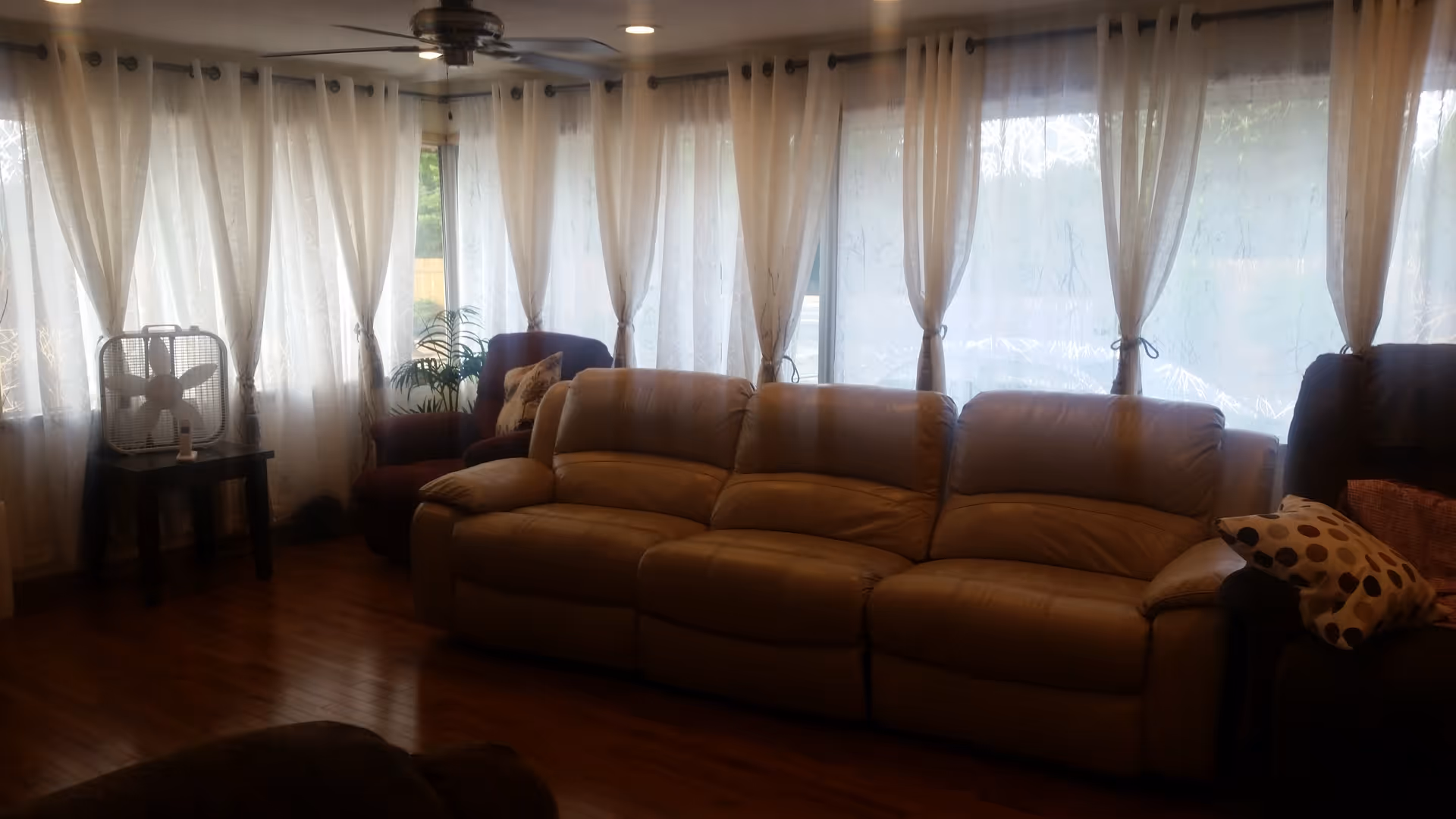 A cozy living room with a large beige leather sofa, two armchairs with patterned pillows, sheer white curtains covering multiple windows, a wooden floor, a small table with a box fan, and a ceiling fan above.