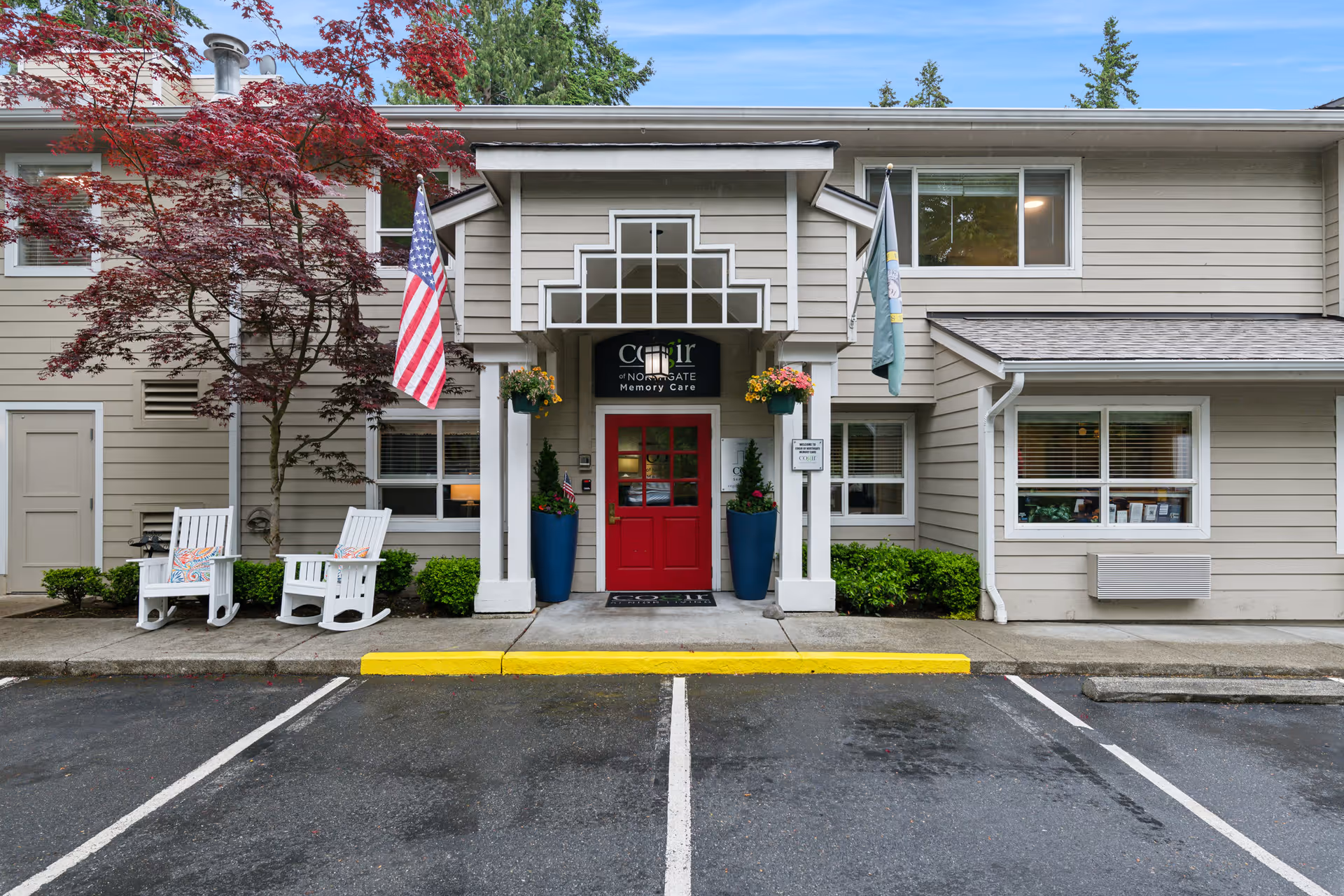 Front entrance of Cogir of Northgate senior living facility with a red door, two white rocking chairs with cushions on the left, American and another flag on either side of the entrance, potted plants, and a tree with red leaves. The building exterior is beige with white trim and windows.