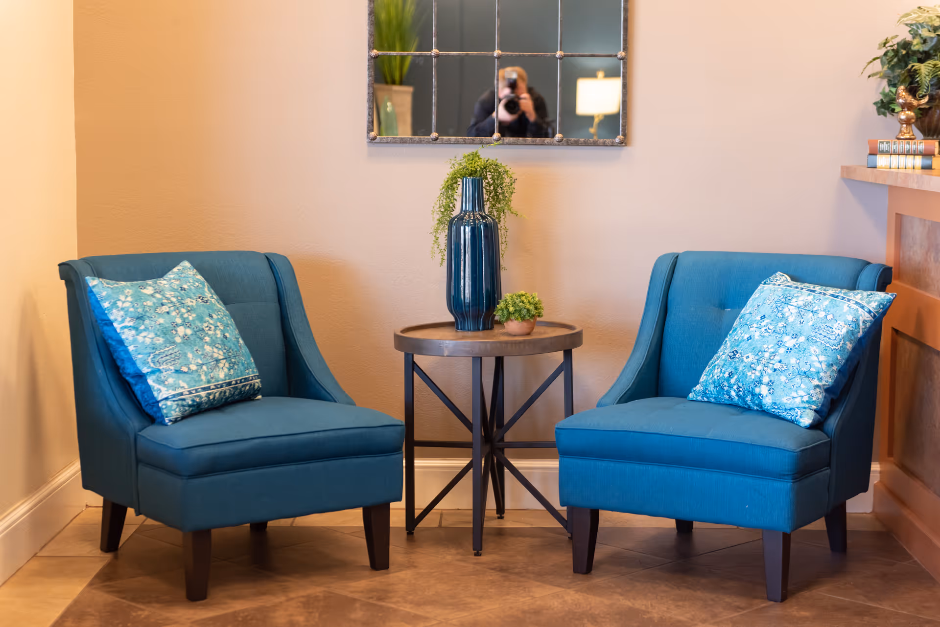 Two blue upholstered armchairs with patterned blue cushions are positioned on either side of a small round wooden table. On the table, there is a tall blue vase with green foliage and a small potted plant. A decorative mirror with a grid pattern hangs on the beige wall behind the chairs. Part of a wooden cabinet with books and a plant is visible on the right side.