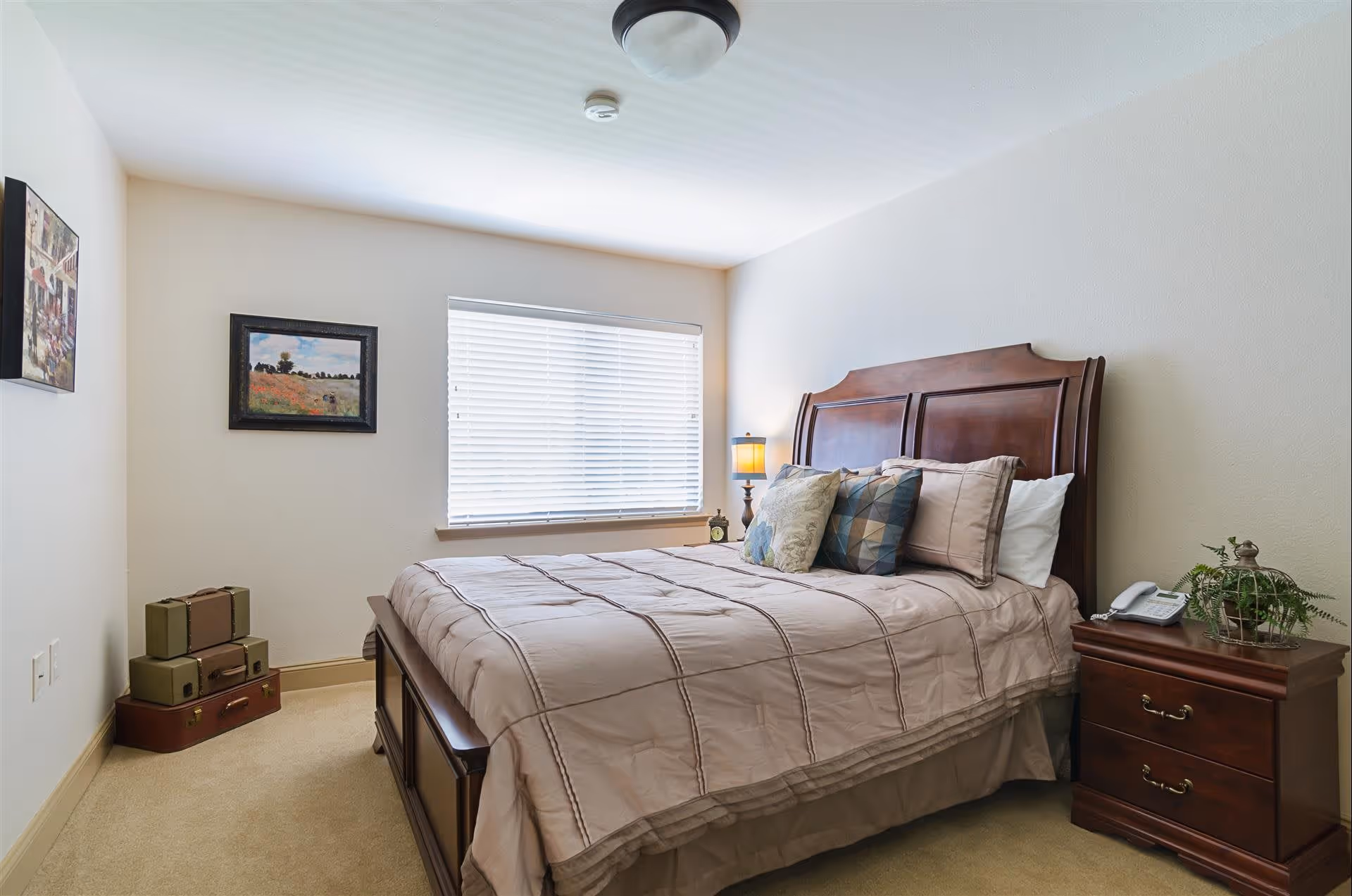 A bedroom with a wooden bed frame and headboard, neatly made with beige bedding and several decorative pillows. There is a wooden nightstand with a lamp, a telephone, and a small plant. A window with white blinds lets in natural light. On the wall, there are two framed pictures, and in the corner, there are stacked vintage suitcases.