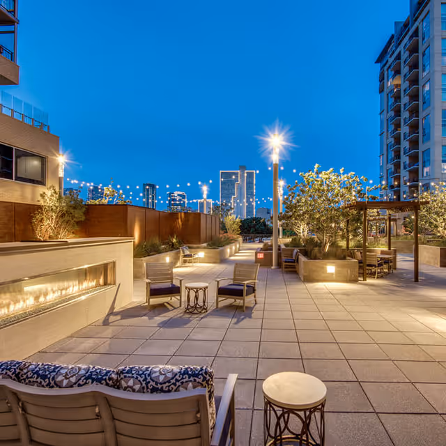 Outdoor patio area at dusk with modern seating arrangements including cushioned chairs and small tables, a long built-in fireplace on the left, string lights overhead, potted plants, and city buildings visible in the background under a clear blue sky.