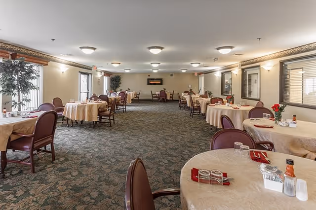 A spacious dining room with multiple round tables covered with beige tablecloths, each set with glasses, napkins, and condiments. The room has patterned carpet flooring, beige walls with decorative trim near the ceiling, and several windows with blinds. There are chairs with maroon cushions around each table and some potted plants near the windows.
